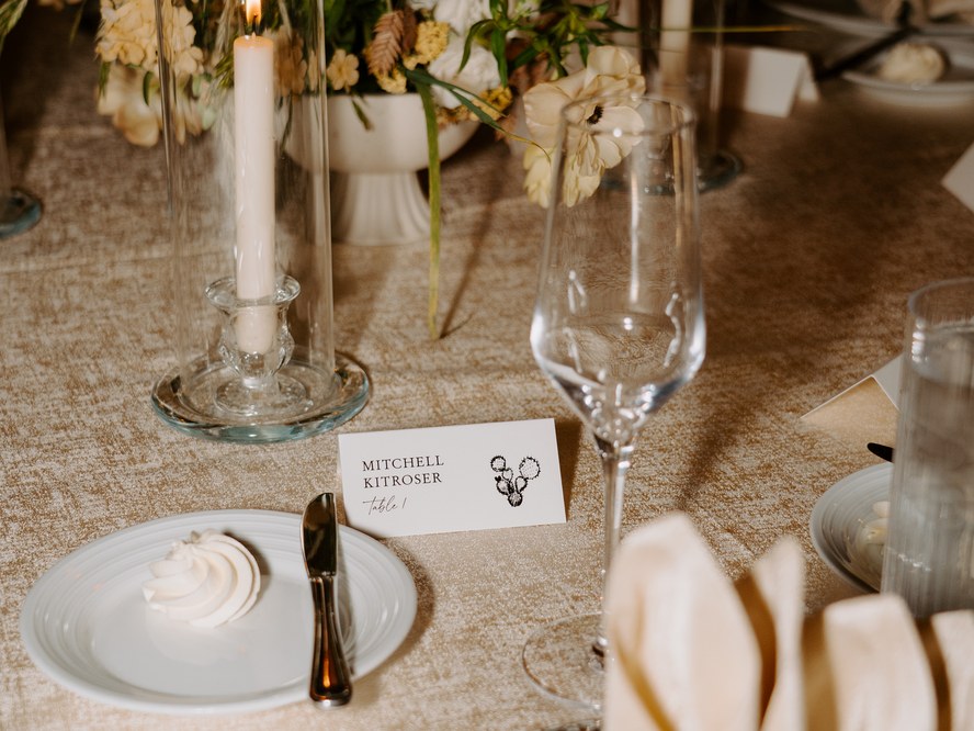 Fall wedding reception place setting with a printed place card, taper candle, autumn florals and a champagne flute on textured gold linen, designed by Be In Love Designs