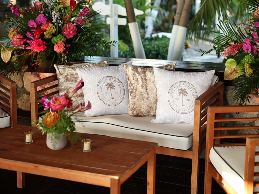 Outdoor event lounge with custom crest-embroidered pillows, tropical florals, and a candlelit cocktail table.